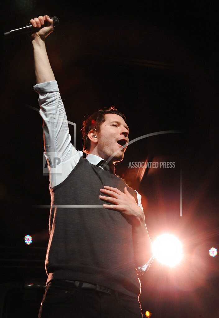 Adam Young holding a microphone in the air on a stage.