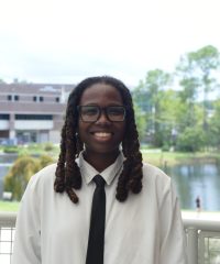 Person with glasses, shoulder length dark hair. white shirt and black tie. Outside background.
