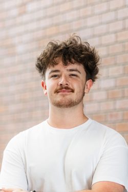 Collin has medium length brown hair and a mustache, and is standing near a brick wall.