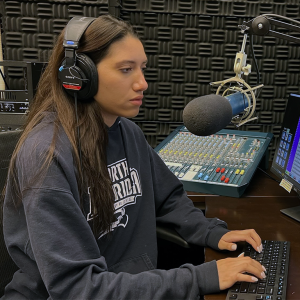 Beija, in a blue sweatshirt and wearing headphones, sits in front of the microphone